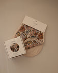 A set of mushie Organic Cotton Muslin Burp Cloth 2-Pack, floral-patterned baby bibs in beige packaging, and a matching boxed pacifier clip are arranged on a neutral beige surface.