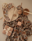 A mushie Organic Cotton Muslin Swaddle Blanket, mauve baby bowl with white spoon, beige pacifier, teether, pink silicone stacking toy, and folded cloth tied with ribbon are arranged on a cream surface.