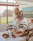 A woman holding a baby stands by a dining table set for a meal with mushie Round Dinnerware Plates, autumn-themed decor, and glasses in a bright room with large windows overlooking nature.