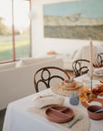 A sunlit dining table set for a meal features ceramic dinnerware and mushie Silicone Feeding Spoons beside a tall pink candle and autumnal decor, with wooden chairs and cozy living room landscape art in the background.