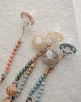 Four mushie Silicone Pacifier Clip | Eva sets, each featuring scalloped-edge pacifiers in soft pastels and food-grade silicone clips with wooden and silicone beads, are displayed on a white textured surface.