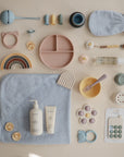 A neatly arranged flat lay of pastel baby items—including a mushie Silicone Pacifier Clip (Eva), towel, mitt, plates, utensils, toys, bottle, teethers, and baby care products—displayed on a beige background.