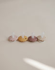 The FRIGG Daisy Silicone Pacifier 2-Pack by FRIGG features medical-grade silicone pacifiers in brown, pink, mustard yellow, and beige with soft white nipples, arranged on a light surface to ensure your baby's comfort.