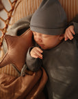 A baby sleeps on a brown cushion, holding the mushie Star Lovey Blanket, nestled in a wicker basket with warm lighting and a gray ribbed outfit and hat.