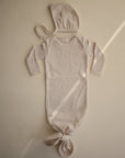 A mushie Ribbed Baby Bonnet in beige is paired with a matching newborn knotted sleeper gown, both displayed flat on a neutral background, softly lit with a sunbeam highlighting the organic cotton fabric.