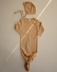 A beige, long-sleeved baby gown with a knotted bottom lies on a cream background. Above it is the mushie Ribbed Baby Bonnet with ties, crafted from organic cotton. Sunlight casts diagonal lines across the scene.