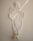 A mushie Ribbed Baby Bonnet in cream is laid flat with a matching long-sleeve knotted gown on a light beige surface, softly lit across the scene.