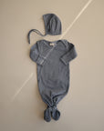 A soft, long-sleeve blue baby gown with a knotted bottom is laid flat on a neutral background. Above the gown is the mushie Ribbed Baby Bonnet in matching blue with ties, creating a cozy newborn set.