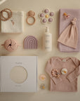 Flat lay of pastel baby items including mushie’s pink bodysuit, hat, muslin cloths, teething toys, pacifier clip, silicone rainbow, lotion bottle, snack container, and a boxed mushie Baby Wrap—ideal for comfortable baby wearing.