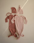 Two mushie Ribbed Baby Bonnets are arranged above two knotted baby gowns, one dusty rose and one light pink, all displayed side by side on a neutral background.