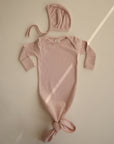 A pale pink long-sleeve baby gown with a knotted bottom is laid flat on a neutral background. Above it rests the mushie Ribbed Baby Bonnet, made from organic cotton. Diagonal light streaks add a soft touch to the image.