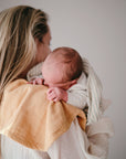 A woman with long blonde hair holds a newborn over her shoulder, using the mushie Organic Cotton Muslin Burp Cloth 2-Pack. The baby rests its head on the soft cloth, wrapped in a light blanket against a neutral background.