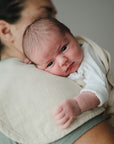 A newborn in a white onesie rests on an adult’s shoulder, supported by mushie Organic Cotton Muslin Burp Cloth 2-Pack draped nearby. The adult wears a green shirt; the softly blurred background highlights the cozy scene.