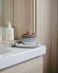 A stack of mushie Bath Boats in pastel silicone sits on a washcloth by the bathroom sink, creating a playful scene with a bottle of soap, a soft toy cube, and light wood cabinets in the background.