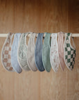 Nine mushie Muslin Bibs in assorted colors and patterns—checks, paisley, solids, and florals—are neatly displayed on a wooden rod with a wood background.