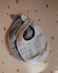 Two mushie Muslin Bibs—one gray and one light beige—are placed side by side on a beige textured surface with small brown rainbow patterns. Both bibs have snap closures at the neck.
