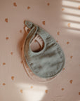 Two mushie Muslin Bibs, one in sage green and one in light pink, are stacked on a peach fabric with an orange rainbow pattern. Window-blind light creates soft shadows across the scene.