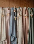A row of soft, pastel baby blankets hangs on a wooden rod, each adorned with the mushie Silicone Pacifier Clip | Eva and matching pacifier. A wooden background highlights these charming mushie accessories.