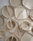 Beige mushie Round Dinnerware Bowls made from BPA-free plastic are arranged with matching plates and utensils in an overlapping pattern on a white surface, some featuring coordinating forks or spoons inside.