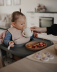 A baby in a high chair practices fine motor skills with the mushie Flower Press Toy. Wearing a bib, the baby sits before cut fruit and a spoon in a bright kitchen and living area.