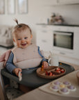 A smiling baby with a ponytail sits in a highchair, enjoying snacks from the mushie Silicone Suction Plate. The scene features a bright, modern kitchen designed for feeding time.
