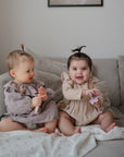 Two babies sit on a beige sofa. The baby on the right, smiling in a beige outfit, holds a soft toy, while part of the sofa is covered with the mushie Organic Cotton Muslin Swaddle Blanket.
