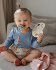 A smiling baby with a FRIGG Daisy Natural Rubber Pacifier sits on a bed holding a toy paint palette, wearing a blue long-sleeve shirt and patterned shorts, with a small ponytail and a pink blanket nearby.