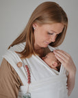 A red-haired woman lovingly cradles her baby in a mushie Baby Wrap made from organic cotton. Wearing a beige shirt against a plain background, she enjoys the comfort and closeness the wrap provides.