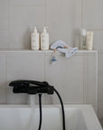 A modern bathroom with a white tub, black faucet, beige tiled wall, water running. On the edge: skincare bottles, washcloth, Mushie Organic Cotton Bath Mitt for sensitive skin, baby pacifier with clip, and a small bottle.