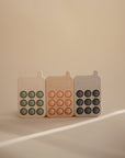 Three mushie Phone Press Toys—pastel-colored silicone fidgets shaped like vintage cell phones—stand side by side on a neutral surface, inviting sensory play in soft light with minimal background shadows.