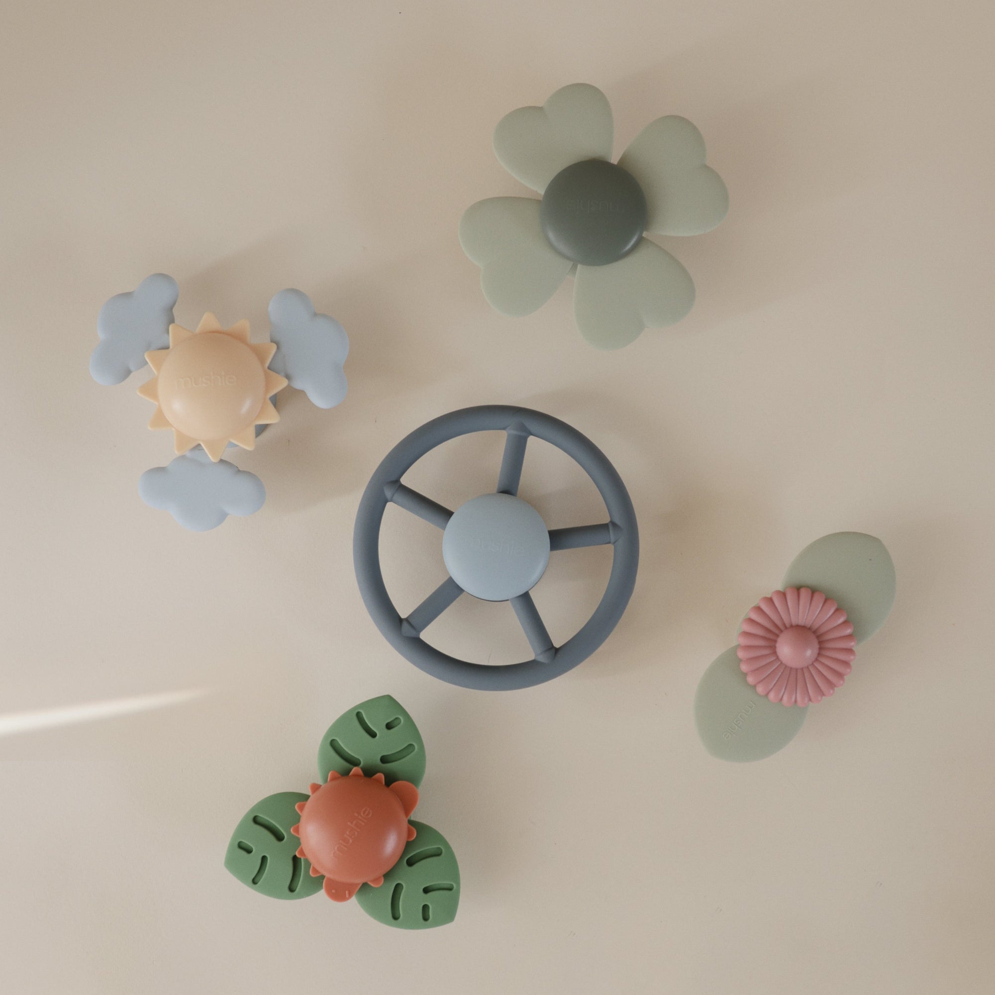 Five mushie Suction Spinner Toys in pastel hues—shaped like a wheel, flower, cloud, and leaves—are crafted from food-grade silicone and arranged on a light beige background for a delightful sensory experience.