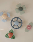 Five mushie Suction Spinner Toys in pastel hues—shaped like a wheel, flower, cloud, and leaves—are crafted from food-grade silicone and arranged on a light beige background for a delightful sensory experience.