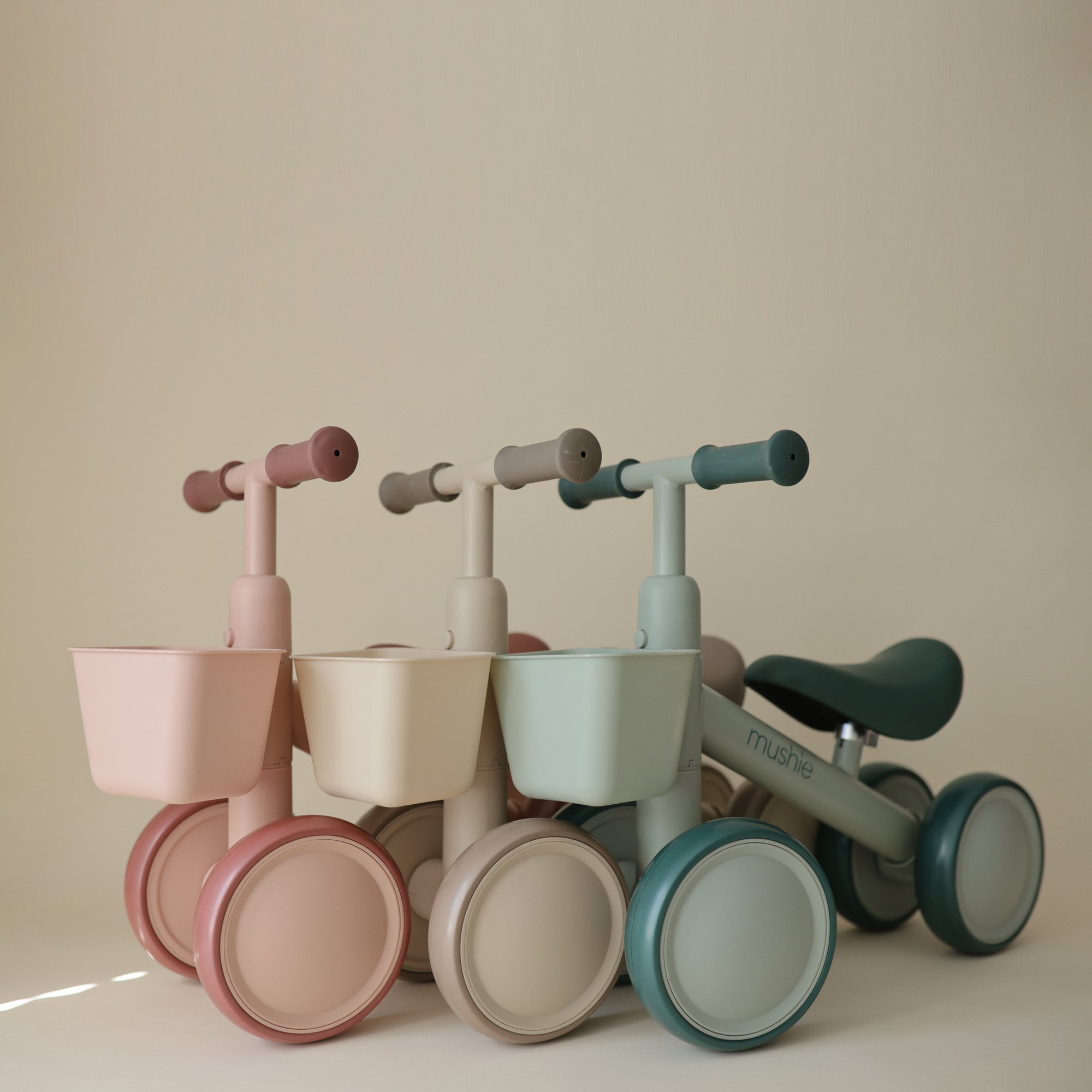 Three mushie Mini Toddler Bikes in pastel colors are lined up against a beige background.