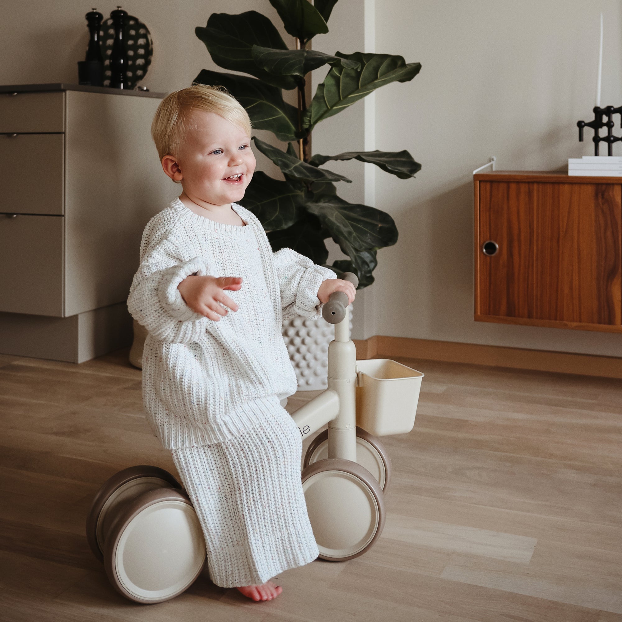 Smiling toddler rides a beige mushie Mini Toddler Bike indoors near plants and furniture.