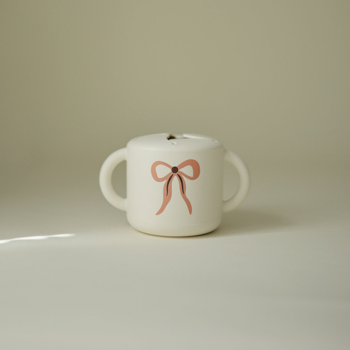 Mushie Snack Cup: spill-proof, white, two handles, pink bow design, plain background.