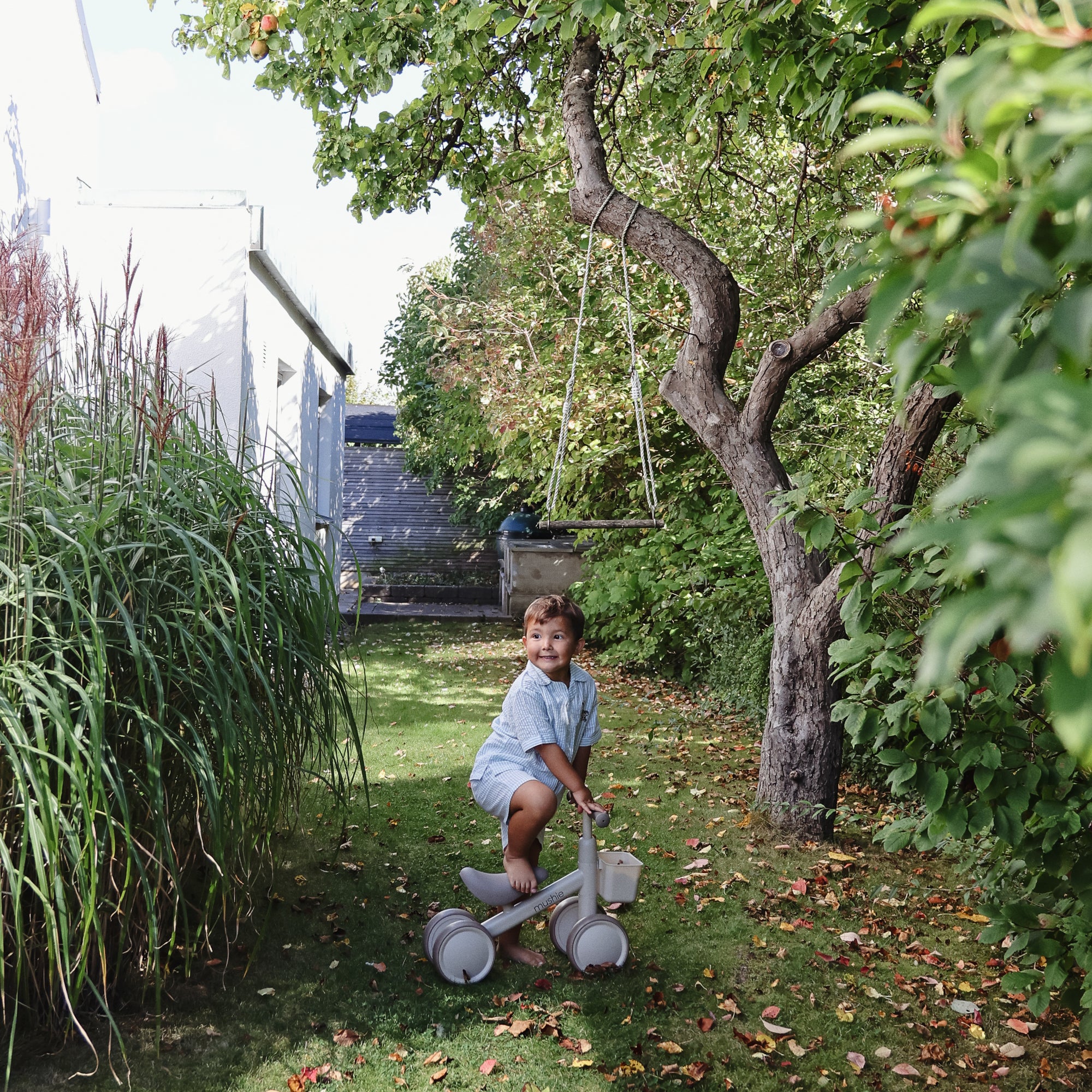 A toddler rides the mushie Mini Toddler Bike on a grassy path under a tree.