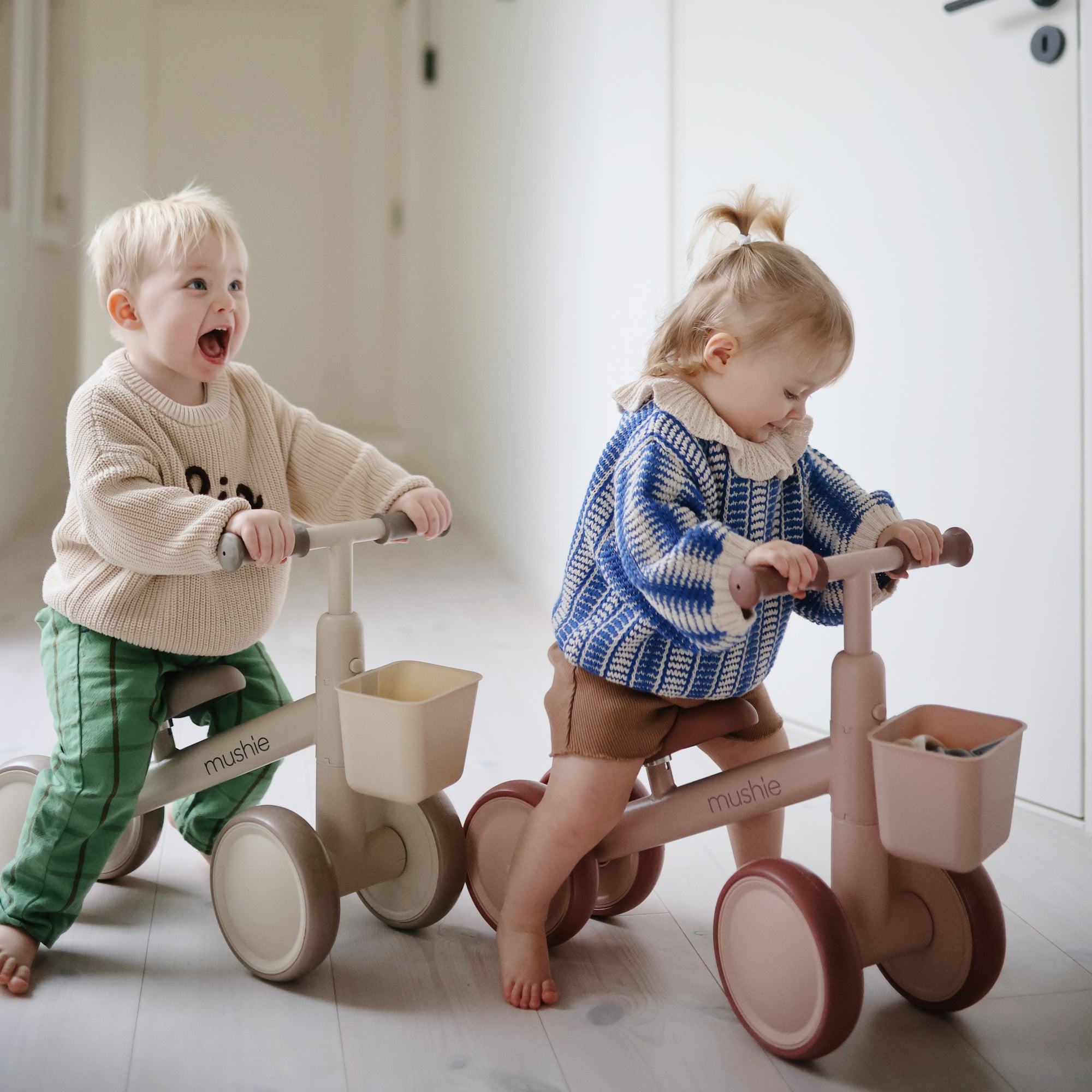 Two toddlers ride their mushie Mini Toddler Bike indoors—one smiles, the other looks ahead.