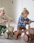 Two toddlers ride their mushie Mini Toddler Bike indoors—one smiles, the other looks ahead.