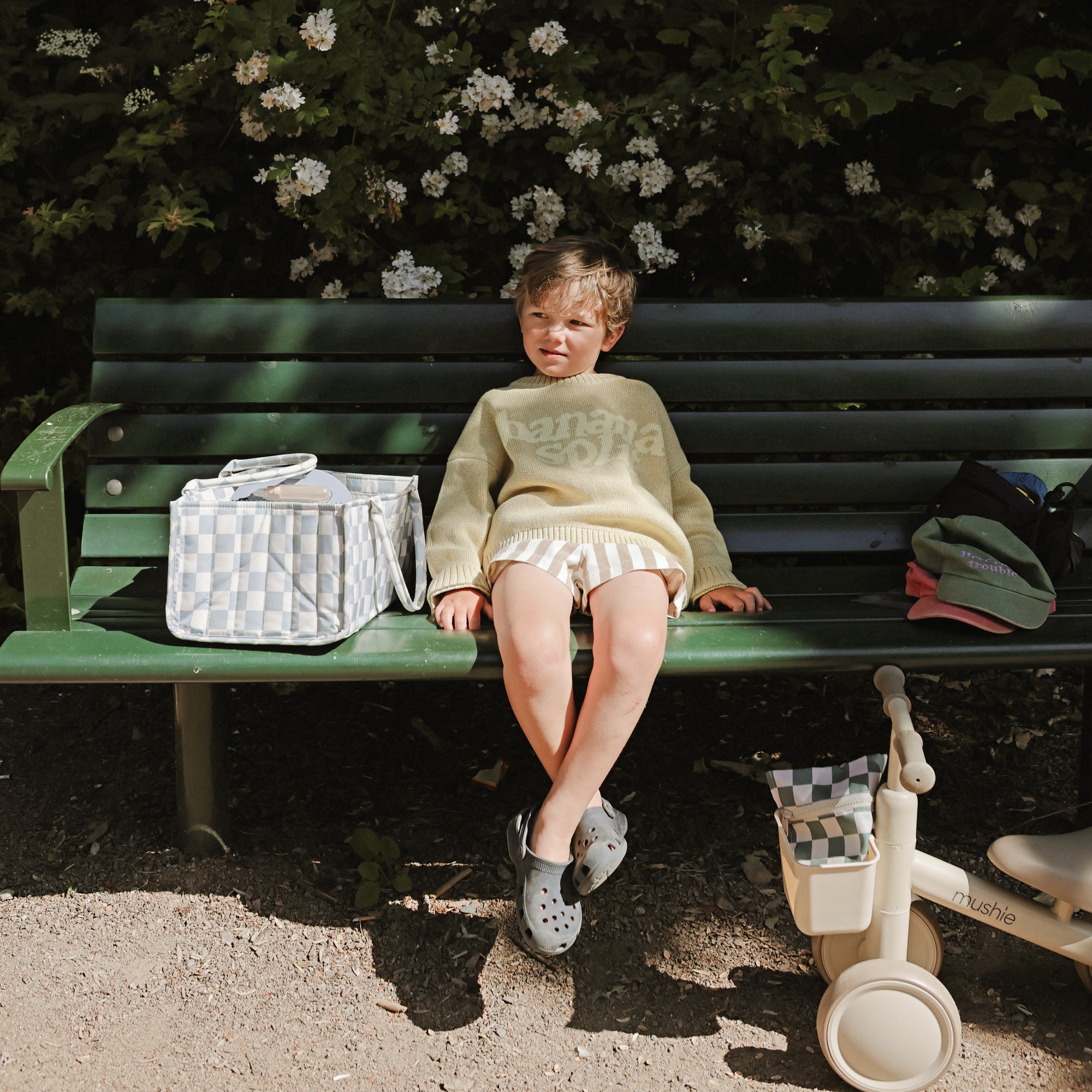 Child sits by green bench, with mushie Diaper Caddy and tricycle nearby in the sunlight.