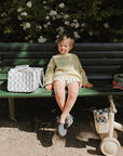 Child sits by green bench, with mushie Diaper Caddy and tricycle nearby in the sunlight.