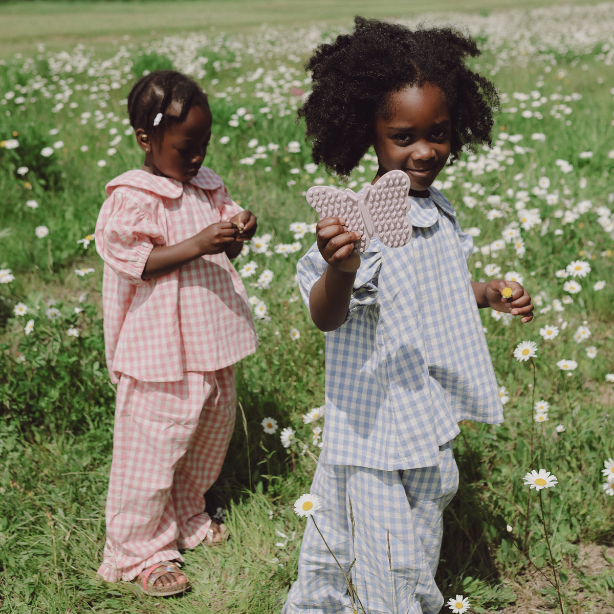 Two kids in gingham pick flowers; one holds a mushie Silicone Sensory Toy shaped like a butterfly.