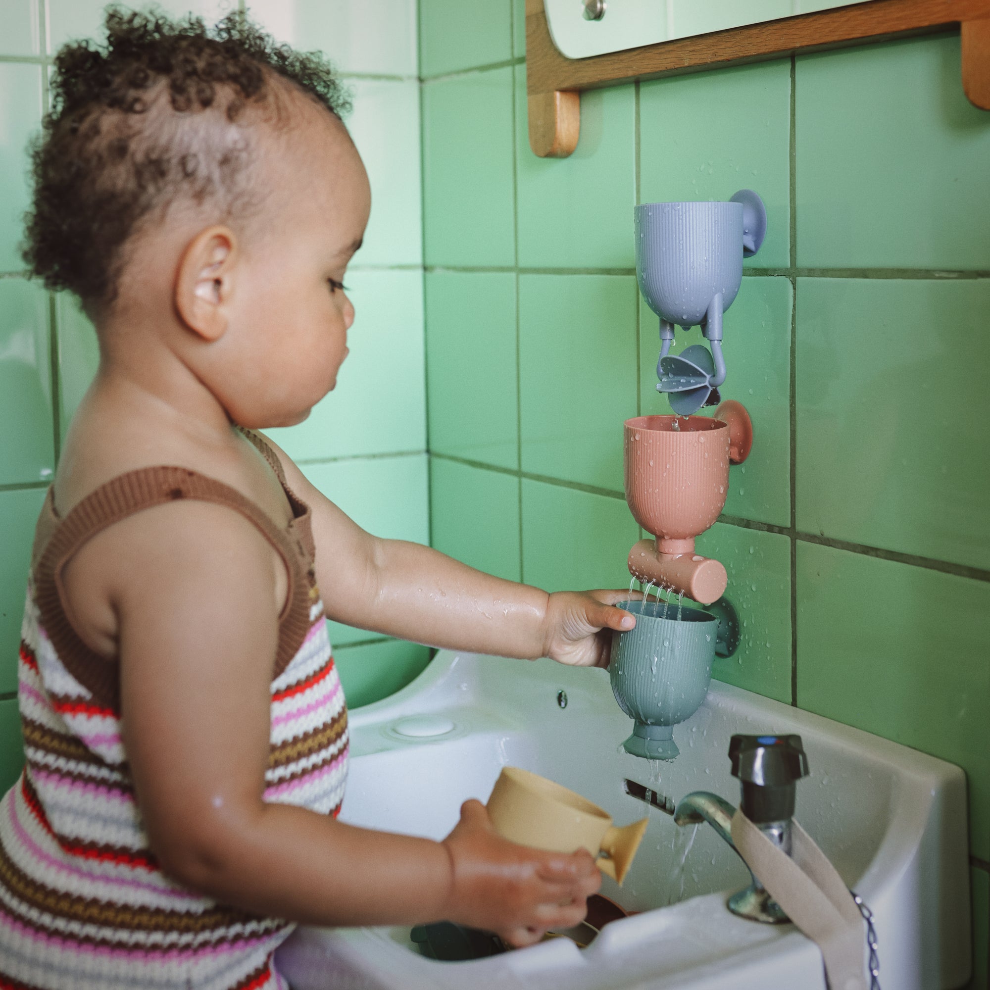 A toddler explores with mushie's Pour and Play Bath Set in a green-tiled bathroom sink.
