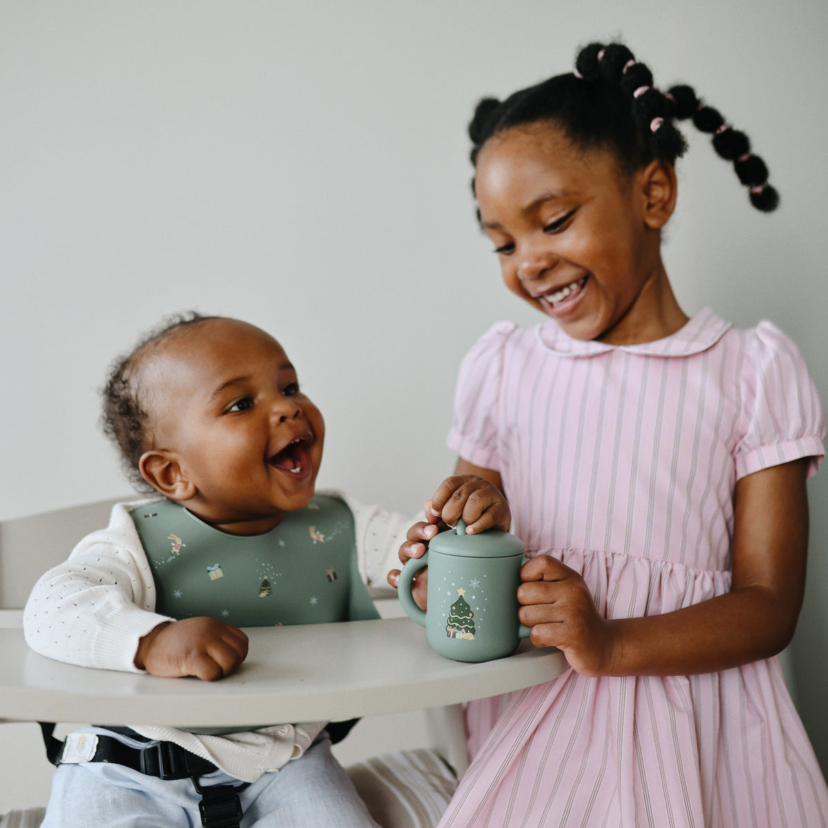 Girl hands a cup to a smiling baby in a high chair wearing mushie Silicone Baby Bib.