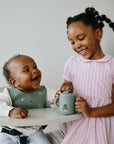 Girl hands a cup to a smiling baby in a high chair wearing mushie Silicone Baby Bib.