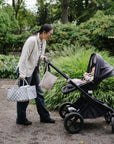 A woman pushes a stroller with a patterned diaper bag and a mushie Water Resistant Wet Bag.