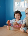 Two kids play with the mushie Mold Free Bath Play Set at a wooden table in a blue room.