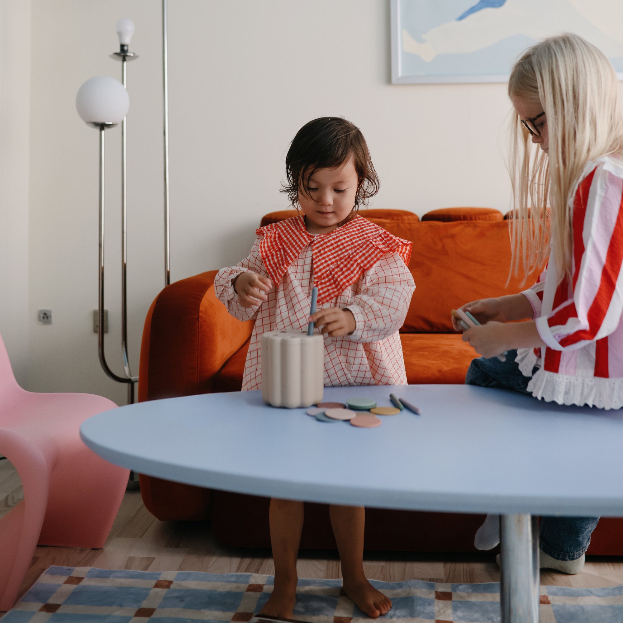 Two young children play at a light blue table with the mushie Coin &amp; Tube Sorting Set, developing fine motor skills as they sort and arrange colorful pieces. An orange sofa and a modern lamp are seen in the cozy living room background.