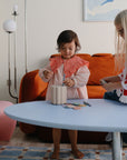 Two young children play at a light blue table with the mushie Coin & Tube Sorting Set, developing fine motor skills as they sort and arrange colorful pieces. An orange sofa and a modern lamp are seen in the cozy living room background.
