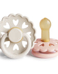 Two baby Frigg Andersen Rubber pacifiers, one Cream with heart designs and the other Blush on a white background.
