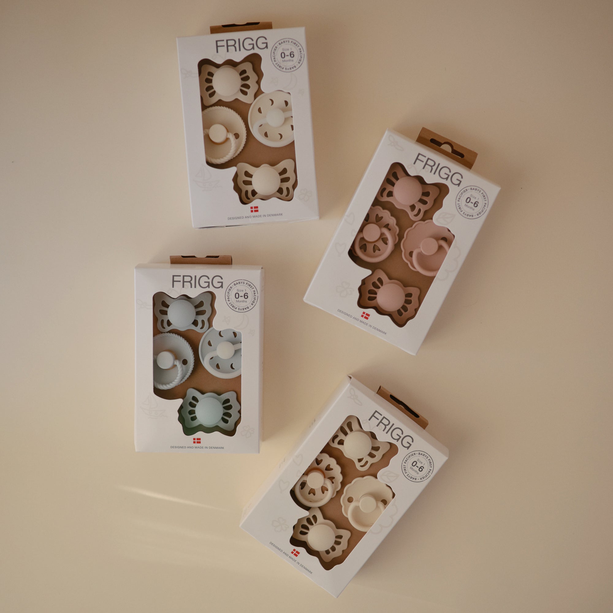 Four boxes of FRIGG Baby's First Pacifier Floral Heart (Blush) 4-Pack, made in Denmark, are arranged on a light surface. Each box displays two pastel pacifiers through a clear window, highlighting the charming floral heart design.